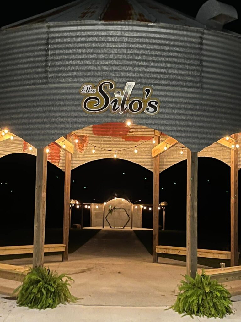 A metal barn-like structure labeled "The Silo's" is lit by string lights at night, with wooden beams, ferns at the entrance, and a decorated arch visible in the background.