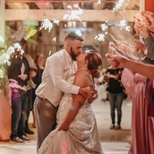 A bride and groom share a kiss while dancing, surrounded by guests holding sparklers. The scene is festive and joyful, with everyone celebrating the couple’s wedding.