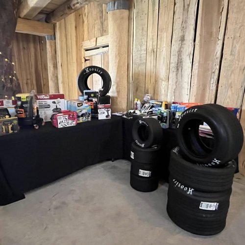 A table displays various automotive products and tools next to stacks of Hoosier racing tires inside a rustic wooden building. A person stands partially visible to the left.