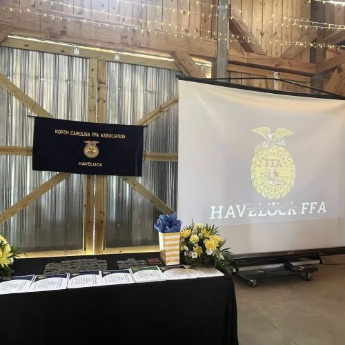 A decorated event setup features a table with certificates and flower arrangements, a screen displaying the Havelock FFA emblem, and a North Carolina FFA Association banner inside a rustic wooden venue.